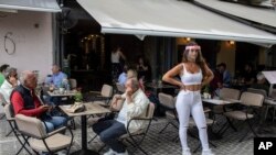A cafe-restaurant staff stands outside as people drink coffee in Monastiraki district of Athens, on Monday, May 25, 2020. Greece restarted regular ferry services to its islands Monday, and cafes and restaurants were also back open for business as…